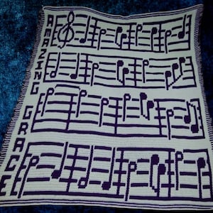 Può includere: Una coperta bianca all'uncinetto con note musicali viola e le parole "AMAZING GRACE" sul lato.