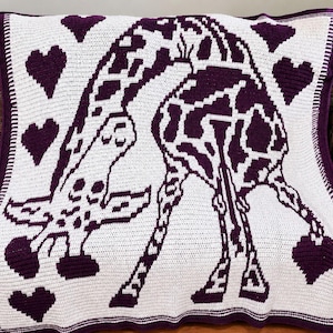 Puede incluir: Una manta de ganchillo rectangular con fondo blanco y un gráfico grande de una jirafa de color morado oscuro en el centro. La jirafa está rodeada por varias formas de corazón de color morado oscuro. La manta tiene un borde morado.