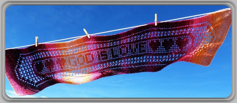 May include: A rectangular crochet scarf featuring a gradient of orange, pink, and purple yarn. The scarf displays the text "GOD IS LOVE" in a block-style pattern, accented by heart shapes, and is suspended horizontally by three wooden clothespins.