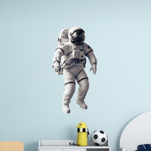 Può includere: Un adesivo di un astronauta in scala di grigi fluttua nella cameretta di un bambino. La stanza ha pareti azzurre, due piccole sedie di legno, un armadio bianco e un letto bianco. Un'anatra giocattolo gialla e un pallone da calcio sono sull'armadio.