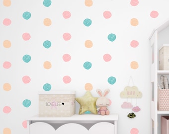 May include: A white bookshelf with three shelves, each containing a storage box. The top shelf has a beige box with a house design, the middle shelf has a pink box with a geometric pattern and a birdhouse, and the bottom shelf has a pink box with a cloud cutout. The bookshelf is against a white wall with a pattern of pastel pink, peach, and turquoise circles.