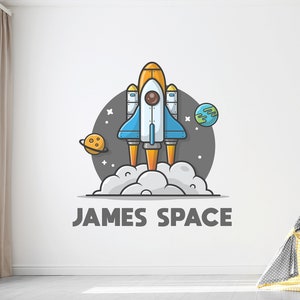Peut inclure: Un mur blanc avec une illustration de dessin animé d'une fusée décollant. La fusée est jaune, bleue et orange. Le texte "JAMES SPACE" est sous la fusée.