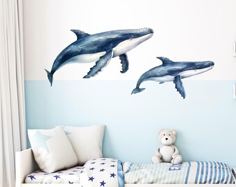 May include: Two watercolor whale wall decals in shades of blue, one larger than the other, on a light blue wall above a white bed with a blue and white striped bedspread and a blue star patterned blanket.