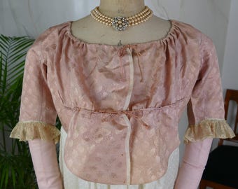 Puede incluir: Una chaqueta de brocado rosa con ribete de encaje blanco en las mangas. La chaqueta está atada a la cintura con una cinta. La chaqueta se lleva sobre un vestido blanco con estampado de lunares. La modelo lleva un collar de perlas con un broche de plata y pedrería.