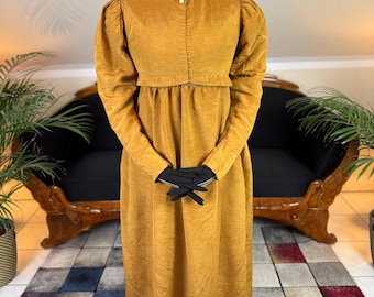 May include: Mustard-colored vintage dress with a high collar and long sleeves. The dress features a fitted bodice, a gathered waist, and a floor-length skirt. A decorative brooch adorns the collar. Black gloves are worn on the hands.