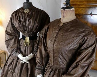 May include: Two brown vintage dresses on dress forms. The dress on the left has a fitted bodice and a full skirt. The dress on the right has a more fitted silhouette. Both dresses have long sleeves and are made of a textured fabric.