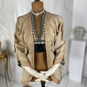 Puede incluir: Una chaqueta beige con ribetes dorados y borlas. La chaqueta tiene un cuello de terciopelo y está decorada con botones de imitación de perlas.
