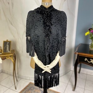 May include: A black velvet cape with a floral pattern and fringe trim. The cape is displayed on a mannequin wearing white gloves.