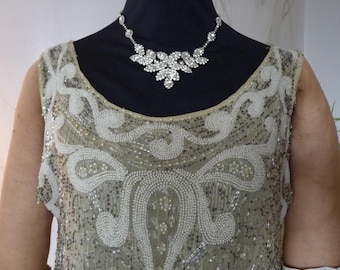 May include: A sleeveless beaded gown with a delicate floral pattern. The gown is a pale beige color and features a sparkling rhinestone necklace.
