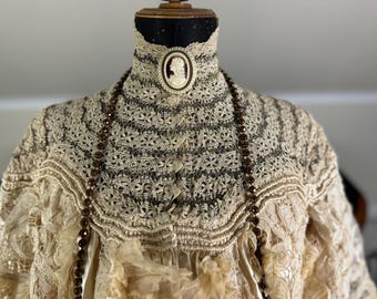 May include: A vintage lace top with a high neckline and a cameo brooch. The top is adorned with a brown beaded necklace.