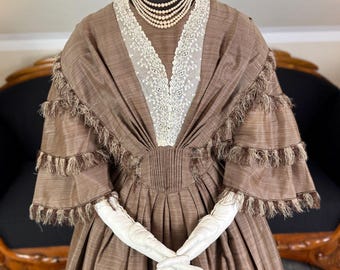 May include: A brown dress with a lace neckline and a shawl collar. The dress has a fitted bodice and a full skirt. The dress is trimmed with brown fringe and velvet ribbon. The dress is being displayed on a mannequin.