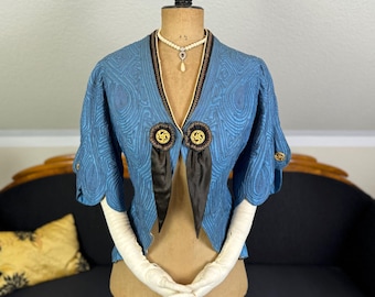 May include: A blue quilted jacket with black trim and a black satin bow. The jacket has a V-neck and two large buttons with gold accents. The jacket is on a mannequin with white gloves.