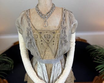 May include: A vintage-style dress on a mannequin. The dress features a beige bodice with intricate embroidery and a layered skirt. A silver necklace with a teardrop pendant adorns the neckline. Long white gloves complete the ensemble, suggesting a historical or formal theme.