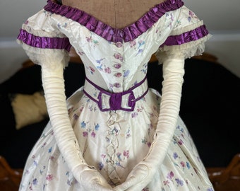 May include: A white silk dress with a floral print and purple trim. The dress has a fitted bodice and a full skirt. The dress is being worn on a mannequin.