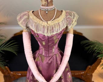 May include: A vintage mauve dress with gold floral accents, displayed on a mannequin. The dress features a fitted bodice, a full skirt, and short, puffed sleeves with lace trim. A pearl necklace and long pink gloves complete the ensemble.