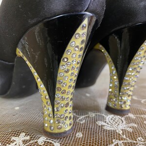 May include: A pair of black satin high heels with a black and gold stiletto heel. The heel is covered in rhinestones.
