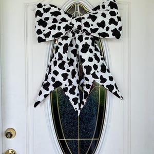 May include: A large decorative bow with a cow print pattern, featuring black spots on a white background. The bow is hanging on a white door with an oval glass window. The bow's design includes long, pointed tails.