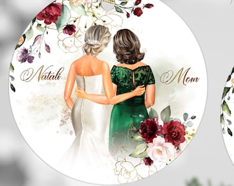 May include: Two round, white, ceramic ornaments with gold ribbon hangers. The first ornament features a watercolour illustration of two women, one in a white wedding dress and the other in a green dress, with the names "Natali" and "Mom" written on either side. The second ornament features a watercolour floral design with the text "As I say "I Do" know that everything I'm today is a reflection of you!"