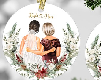 May include: Two round white baubles with gold ribbon hangers. The first bauble features a watercolour illustration of two women, one in a white wedding dress and the other in a burgundy dress, standing back to back with their arms around each other. The text "Kayla Mom" is written in gold script above the women. The second bauble features a watercolour illustration of a floral wreath with white flowers, green leaves, and red berries. The text "Thank you for your support and care I'll always be grateful for the love we share!" is written in gold script inside the wreath.
