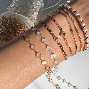 Puede incluir: Se muestra una colección de pulseras de oro y plata en una muñeca. Las pulseras incluyen una pulsera de perlas, una pulsera de cadena y pulseras con dijes en forma de diamante y cuadrado. Se ven carretes de cadena en el fondo.