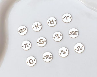 May include: A collection of small, round, gold-colored charms, each engraved with a different zodiac symbol. The charms are scattered on a white surface, with symbols including Aries, Taurus, Gemini, Cancer, Leo, Virgo, Libra, Scorpio, Sagittarius, Capricorn, Aquarius, and Pisces.
