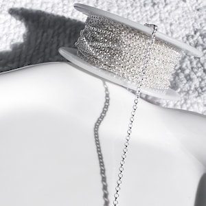 May include: A spool of silver-colored chain sits on a white surface. The chain is wound around a white plastic spool. A length of the chain hangs down, casting a shadow on the surface.