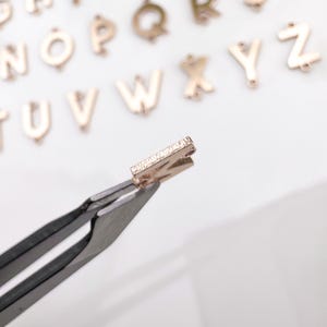 May include: A gold letter charm in the shape of the letter "Z" with a row of small diamonds on the front. The charm is being held by tweezers.