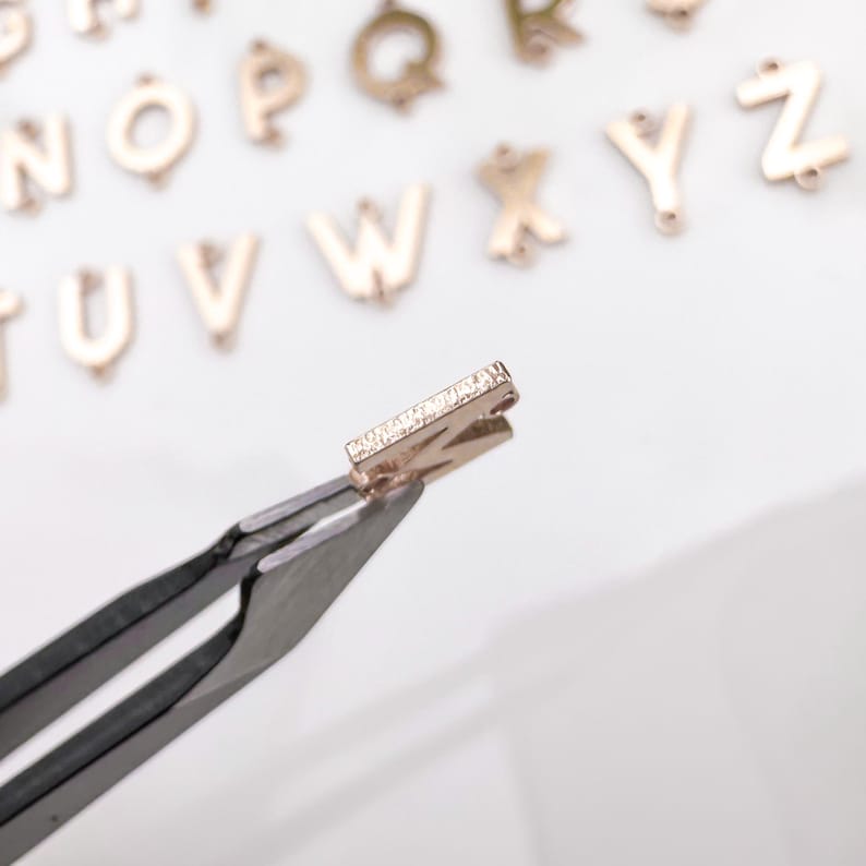 May include: A gold letter charm in the shape of the letter "Z" with a row of small diamonds on the front. The charm is being held by tweezers.
