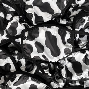 May include: A close-up of a pile of black and white cow print fabric face masks. The masks are made of a soft, silky material and have adjustable ear loops.