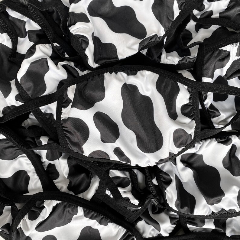 May include: A close-up of a pile of black and white cow print fabric face masks. The masks are made of a soft, silky material and have adjustable ear loops.