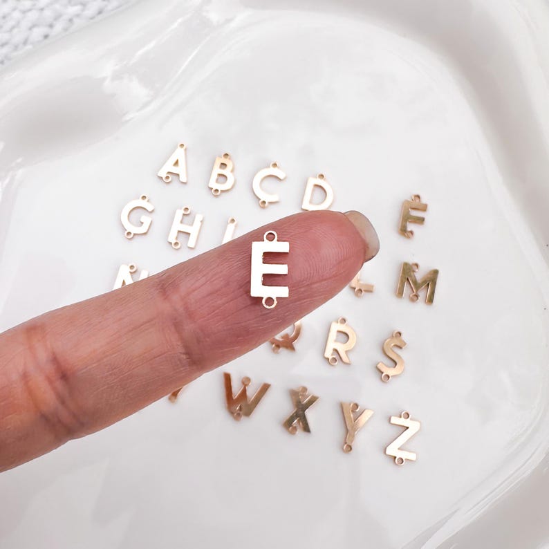 May include: A collection of gold-colored metal alphabet letter charms. The letters are arranged in alphabetical order, with the letter "E" being held by a finger.