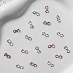 Könnte beinhalten: Eine Ansammlung kleiner, unendlichkeitsförmiger Charms in Gold- und Silbertönen. Die Charms sind auf einer weißen, herzförmigen Oberfläche verstreut, was eine zarte und elegante Ästhetik erzeugt. Ideal für Schmuckherstellung oder Bastelarbeiten.