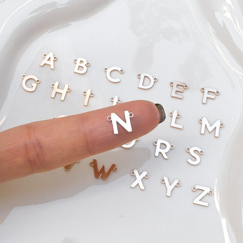 May include: A set of gold-colored metal alphabet letter charms. The letters are arranged in alphabetical order on a white surface. The letter N is being held by a finger.