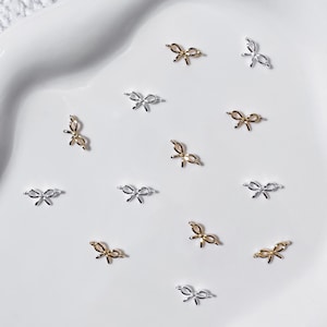 Peut inclure: Une collection de petits breloques pour nail art en forme de nœuds, de tons or et argent. Les breloques sont dispersées sur une surface blanche, créant une esthétique délicate et élégante. Ces accessoires de nail art décoratifs ajoutent une touche de sophistication à toute manucure.