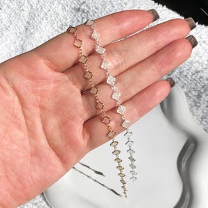 May include: Two delicate necklaces, one gold-toned and one silver-toned, each featuring a repeating pattern of small, square-shaped charms. The necklaces are displayed against a soft, neutral background, highlighting their intricate design and subtle elegance.