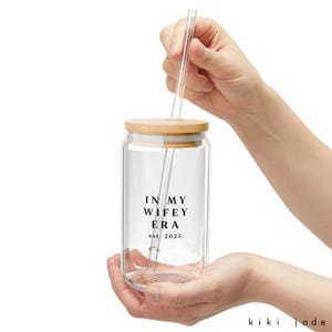 May include: A clear glass tumbler with a bamboo lid and a straw. The tumbler has the text "In My Wife Era est. 2025" printed on it.