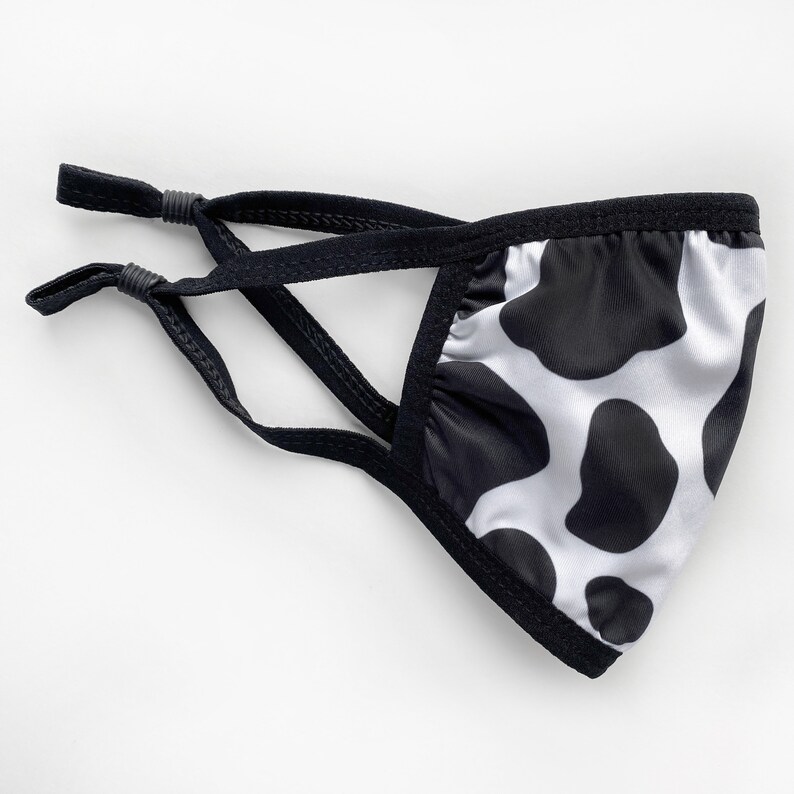 May include: Black and white cow print face mask with adjustable ear loops. The mask is made of a soft, breathable fabric.