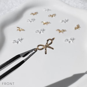 May include: A gold-colored bow-shaped charm is held by tweezers, with several similar charms scattered on a white surface. The charms are small and delicate, with a shiny finish. The word "FRONT" is visible at the bottom of the image.