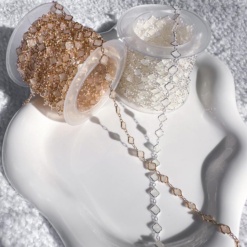May include: Spools of decorative chain in gold and silver tones. The chains feature a repeating geometric pattern of small, square-shaped links. The spools are displayed on a white, wavy-edged plate, with the chains partially unraveled.