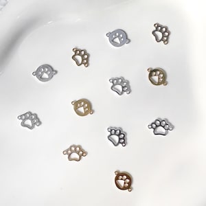 May include: A collection of small, metal paw print charms in silver and gold tones. The charms are scattered on a white surface, showcasing their delicate design and potential for jewelry making or crafting projects.