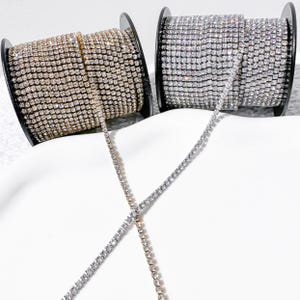 May include: Two spools of rhinestone chain, one gold and one silver, are displayed. The chains are unspooled, crossing each other. The spools are black and sit on a white surface. The rhinestones are small and closely set.