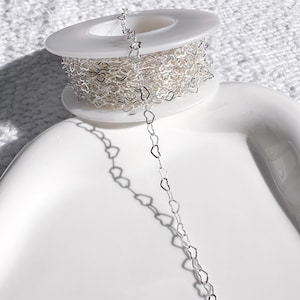 May include: A spool of silver-colored chain with a heart-shaped design. The chain is partially unraveled, revealing the delicate heart links. The spool is white and sits on a white, curved surface. The chain's shadow is visible.