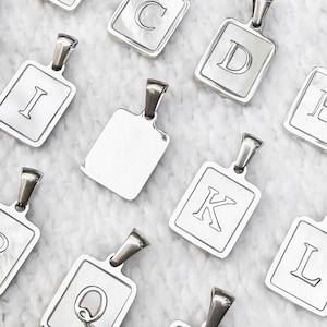 Op de afbeelding: Een set zilverkleurige metalen hangers met verschillende gegraveerde letters. De hangers zijn rechthoekig en hebben een lus aan de bovenkant om aan een ketting of koord te bevestigen.
