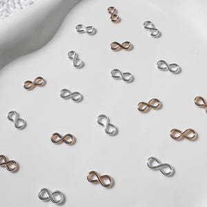 Könnte beinhalten: Eine Ansammlung kleiner Unendlichkeitszeichen-Anhänger in Silber- und Roségoldtönen. Die Charms sind auf einer weißen Oberfläche verstreut und eignen sich für die Schmuckherstellung und das Basteln.