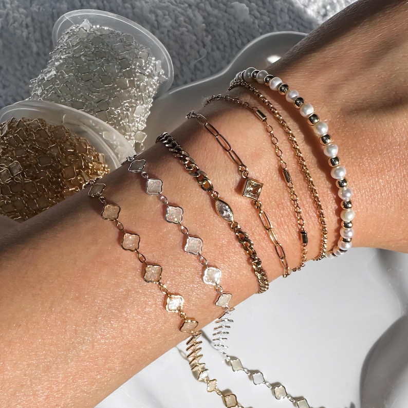 May include: A collection of gold and silver bracelets with various designs, including chain links, diamond-shaped accents, and pearls. Several spools of chain are visible in the background.