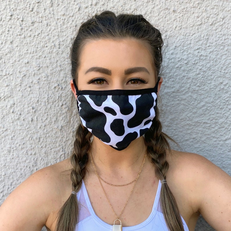 May include: A woman wearing a white and black cow print face mask. The mask has a black elastic band that goes around the ears. The woman is wearing a white tank top and has her hair in two braids.