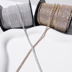 May include: Two spools of rhinestone chain, one in a silver-tone finish and one in a gold-tone finish. Each spool is wrapped with a continuous strand of clear, faceted crystals set in metal, with loose ends crossing over each other in the center.
