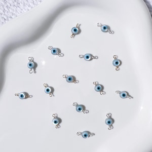 May include: A collection of small, light blue and silver-toned evil eye charms. Each charm features a blue eye design with a black pupil, connected by silver-colored loops. The charms are scattered on a white surface.
