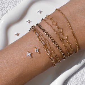 May include: An assortment of gold bracelets and diamond stud earrings. The bracelets display diverse designs, including a chain link, a curb chain, and a delicate chain with diamond-shaped links. The earrings are small, star-shaped studs with clear gemstones.