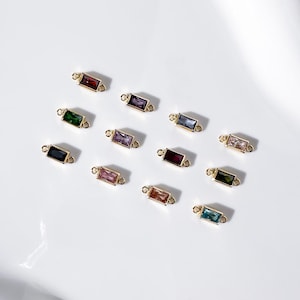 May include: A collection of rectangular gemstone charms in various colors, including red, green, purple, and blue. Each charm is set in a gold-colored metal frame with small loops on each end, suitable for jewelry making.